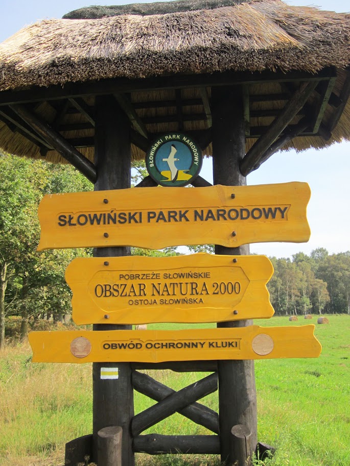 Drewniana tablica informacyjna pod dachem ze strzechy z logo Słowińskiego Parku Narodowego oraz napisami: Słowiński Park Narodowy, Obszar Natura 2000 i Obwód Ochronny Kluki