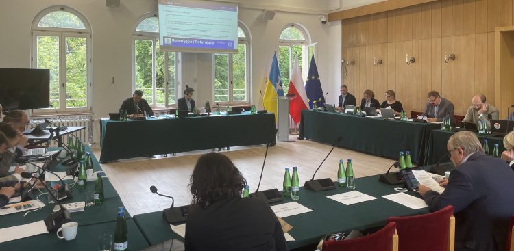 Sesja Rady Miasta Sopotu. Szerokie ujęcie sali obrad, w której radni siedzą przy stołach ułożonych w kształt litery U, przykrytych ciemnozielonymi obrusami. Na stołach znajdują się laptopy, mikrofony konferencyjne, butelki z wodą, dokumenty oraz filiżanki. W tle widoczne są duże, łukowate okna wychodzące, za których widać zieleń, podwieszany pod sufitem ekran z wyświetlaną prezentacją oraz stojące flagi: Ukrainy, Sopotu, Polski i Unii Europejskiej. Kilka osób siedzi tyłem do obiektywu w pierwszym planie, podczas gdy pozostali uczestnicy obrad są zwróceni w stronę stołu prezydialnego