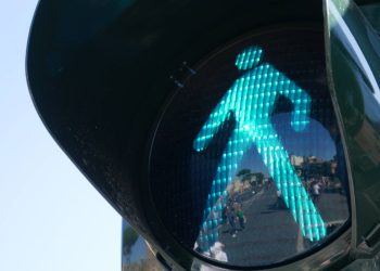 Zbliżenie na zielone światło dla pieszych (tzw. zielony ludzik) na sygnalizatorze. W szklanej powierzchni obudowy odbija się miejska ulica z przechodzącymi ludźmi i budynkami w tle. Po lewej stronie widoczne jest bezchmurne niebo