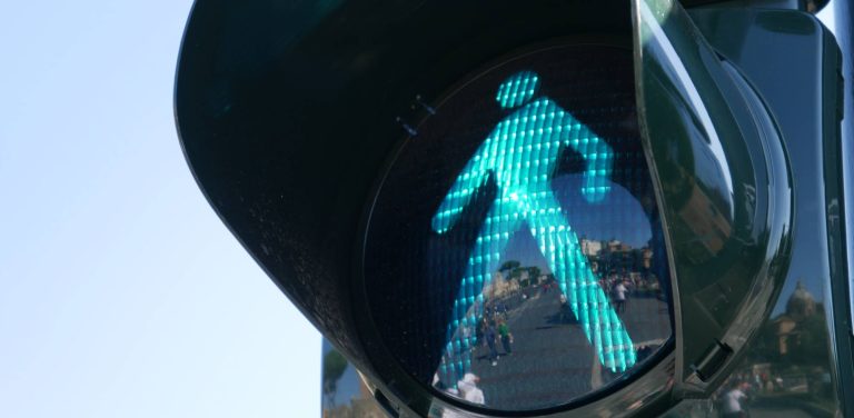 Zbliżenie na zielone światło dla pieszych (tzw. zielony ludzik) na sygnalizatorze. W szklanej powierzchni obudowy odbija się miejska ulica z przechodzącymi ludźmi i budynkami w tle. Po lewej stronie widoczne jest bezchmurne niebo