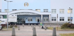 Wejście główne do budynku Szpitala Specjalistycznego im. J. K. Łukowicza w Chojnicach. Nad zadaszonym wejściem widnieje napis z nazwą i adresem placówki, a w tle widać jasnoniebieską elewację z oknami. Na pierwszym planie na chodniku stoją betonowe słupki ochronne oraz zaparkowana hulajnoga elektryczna. Ilustracja informacji dotyczącej zysków wynikających z konsolidacji szpitali w Chojnicach i Człuchowie