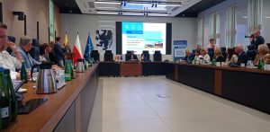 Przy dużym drewnianym stole w kształcie litery U siedzi kilkunastu uczestników. W tle widoczne są flagi Polski, Unii Europejskiej, województwa pomorskiego oraz duży ekran z prezentacją dotyczącą funduszy unijnych. To oficjalne spotkanie samorządowców w sali konferencyjnej w Urzędzie Marszałkowskim Województwa Pomorskiego, gdzie podpisano dokumenty na dofinansowanie unijne w wysokości 75 mln zł dla siedmiu pomorskich gmin na m.in. termomodernizację szkół oraz drogi rowerowe
