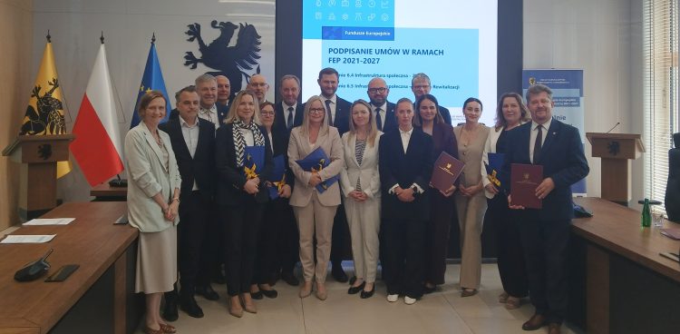 Pamiątkowe zdjęcie grupowe przedstawicieli pomorskich samorządów oraz Urzędu Marszałkowskiego Województwa Pomorskiego. Samorządowcy stoją razem w Sali Herbowej, trzymając w rękach foldery z podpisanymi umowami. W tle widoczny jest duży ekran z napisem „PODPISANIE UMÓW W RAMACH FEP 2021-2027”, czarny gryf na ścianie oraz flagi Polski, Unii Europejskiej i województwa pomorskiego. Dofinansowanie na projekty społeczne na Pomorzu