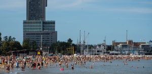 Szeroki kadr przedstawiający zatłoczoną plażę miejską w Gdyni w słoneczny dzień. Na piasku oraz w płytkiej wodzie wypoczywa wielu plażowiczów. W tle, po lewej stronie, wznosi się wysoki, szary wieżowiec Sea Towers oraz sąsiadujący z nim niższy budynek z widocznym czerwonym logo hotelu Courtyard Marriott. Po prawej stronie w tle widoczne są maszty jachtów, żurawie portowe oraz inne zabudowania miejskie pod bezchmurnym, błękitnym niebem