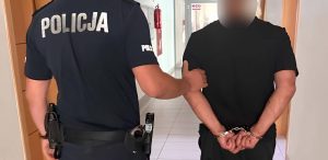 Policjant w granatowym mundurze trzyma za ramię zatrzymanego mężczyznę w czarnym t-shircie, który ma ręce skute kajdankami z tyłu. Twarz zatrzymanego jest zamazana. Mężczyzna został zatrzymany po wtargnięciu do domu żony, do której miał zakaz zbliżania się. Próbował tam podpalić kotłownię oraz oblał łatwopalną cieczą interweniujących policjantów