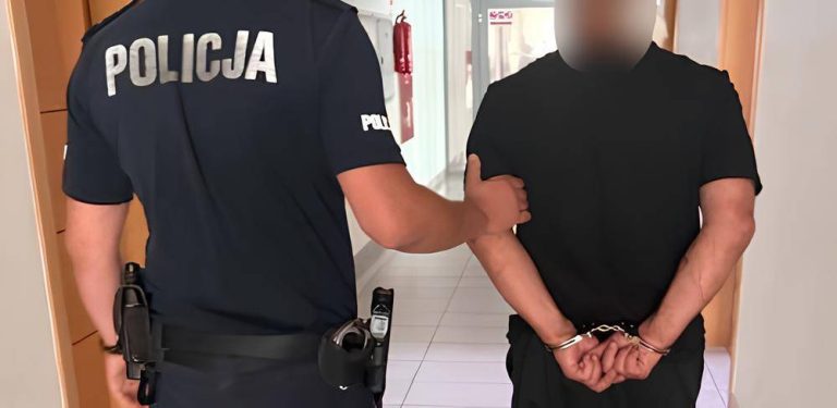 Policjant w granatowym mundurze trzyma za ramię zatrzymanego mężczyznę w czarnym t-shircie, który ma ręce skute kajdankami z tyłu. Twarz zatrzymanego jest zamazana. Mężczyzna został zatrzymany po wtargnięciu do domu żony, do której miał zakaz zbliżania się. Próbował tam podpalić kotłownię oraz oblał łatwopalną cieczą interweniujących policjantów
