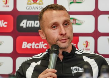 Trener Władysław Łupaszko w czarno-białej bluzie klubowej mówi do mikrofonu podczas konferencji prasowej przed kolejnym meczem w lidze. W tle widoczna jest ścianka z logo sponsorów, miasta Gdańsk oraz klubu Lechia Gdańsk.