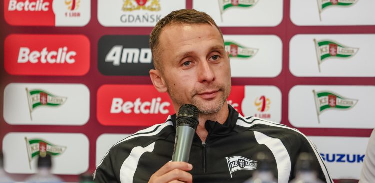 Trener Władysław Łupaszko w czarno-białej bluzie klubowej mówi do mikrofonu podczas konferencji prasowej przed wyjazdowym starciem z Podbeskidziem Bielsko-Biała. W tle widoczna jest ścianka z logo sponsorów, miasta Gdańsk oraz klubu Lechia Gdańsk.