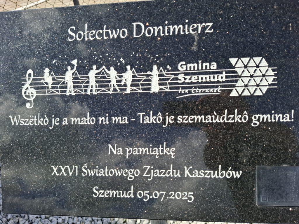 Na zdjęciu tablica z napisem w języku kaszubskim. Tłumaczenie: „Wszystko jest, a mało nie ma - taka jest Szemudzka gmina”.