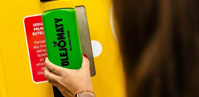 Olejomat w gminie Redzikowo. Kobieca dłoń przykłada zieloną pojemną butelkę z napisem „OLEJOMATY” i żółtą nakrętką do żółtej maszyny do zbiórki oleju spożywczego