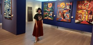 Agnieszka Drączkowską-Gradzik, kuratorka wystawy "Kachu totalny" w Muzeum Miasta Gdyni. Kobieta z krótkimi ciemnymi włosami, ubrana w ciemną koszulkę i długą bordową spódnicę, stoi na tle niebieskiej ścianki w sali muzealnej, na której zawieszone są kolorowe, abstrakcyjne obrazy
