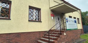 Wejście do budynku Archiwum Państwowego w Słupsku. Schody z poręczami prowadzą do drzwi pod zadaszeniem, na żółtej elewacji widać okna z ozdobnymi kratami, czerwoną tablicę urzędową oraz tablicę informacyjną o unijnym projekcie modernizacji energetycznej