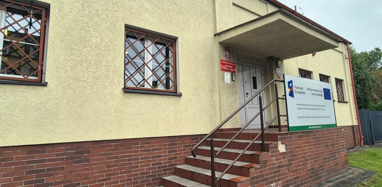 Wejście do budynku Archiwum Państwowego w Słupsku. Schody z poręczami prowadzą do drzwi pod zadaszeniem, na żółtej elewacji widać okna z ozdobnymi kratami, czerwoną tablicę urzędową oraz tablicę informacyjną o unijnym projekcie modernizacji energetycznej