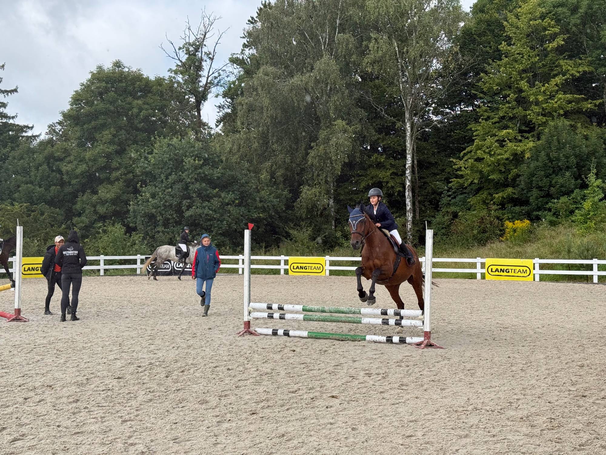 Baltic Cup 2026. Jeździec na kasztanowatym koniu w trakcie skoku przez przeszkodę. W tle widoczni są trenerzy, sędziowie oraz biały płot z żółtymi banerami "Lang Team"