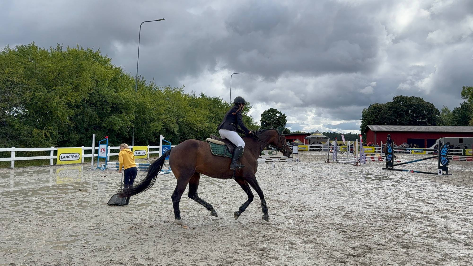 Zawody jeździeckie Baltic Cup 2026. Jeździec na ciemnym koniu porusza się po parkurze w Barnowie. W tle widoczny jest tor przeszkód, osoba w żółtej bluzie porządkująca teren oraz pochmurne niebo