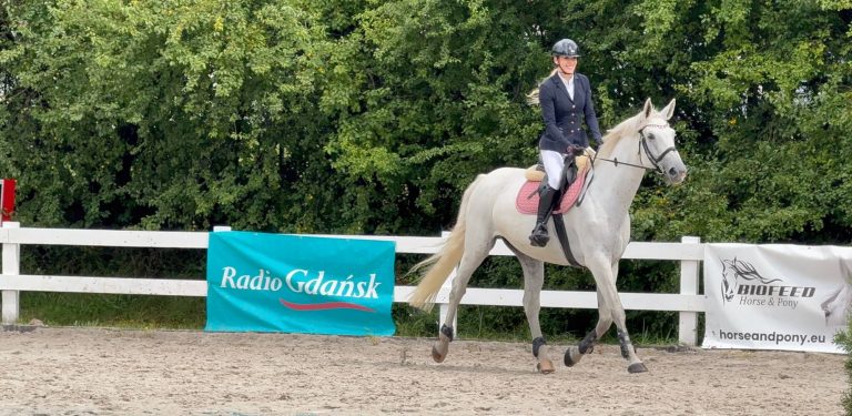 Zawody jeździeckie Baltic Cup 2026. Kobieta w stroju jeździeckim i kasku uśmiecha się, jadąc na jasnoszarym (siwym) koniu. W tle widać biały płot, zielone krzewy oraz banery reklamowe, m.in. z logo "Radio Gdańsk"