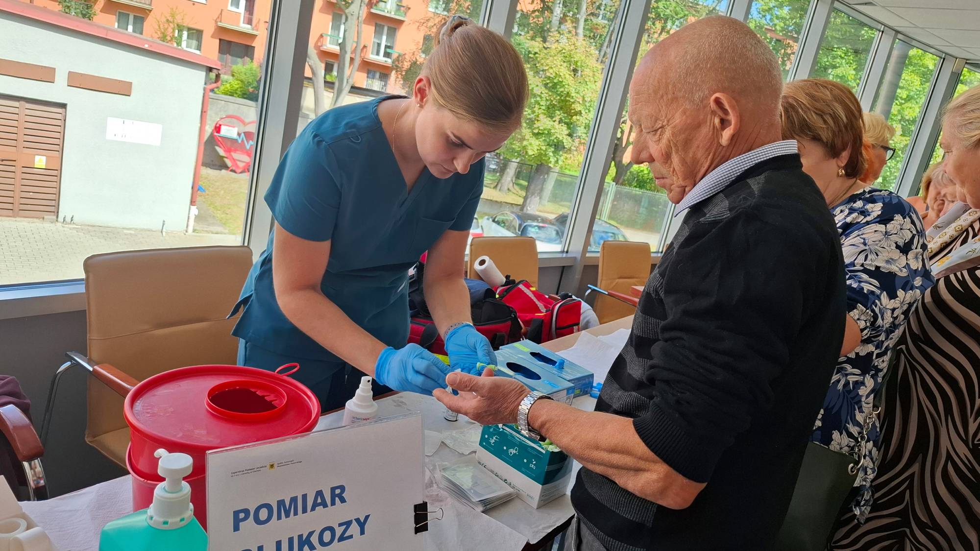 Młoda kobieta w niebieskim stroju medycznym i rękawiczkach pochyla się nad stołem, wykonując pomiar glukozy we krwi u starszego mężczyzny w czarno-szarym swetrze. Na pierwszym planie widoczny jest czerwony pojemnik na odpady medyczne oraz tabliczka z napisem "POMIAR GLUKOZY". W tle przez duże okna widać budynki oraz inne osoby czekające w kolejce