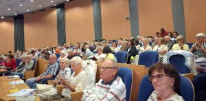 Widok z góry na wypełnioną salę konferencyjną, w której rzędy niebieskich foteli zajmują skupieni seniorzy biorący udział w spotkaniu edukacyjnym