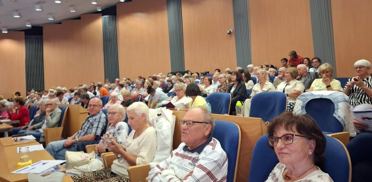 Widok z góry na wypełnioną salę konferencyjną, w której rzędy niebieskich foteli zajmują skupieni seniorzy biorący udział w spotkaniu edukacyjnym