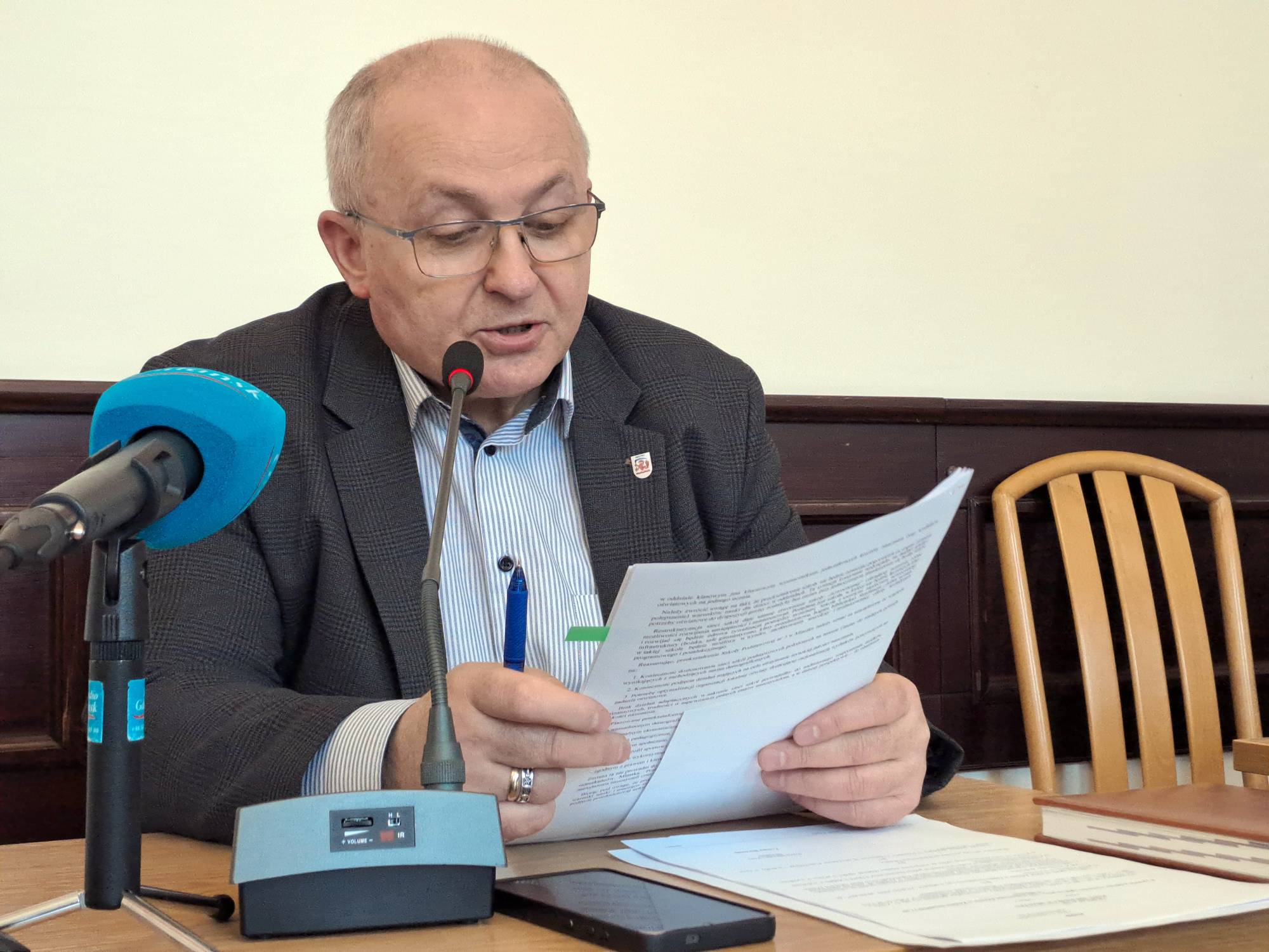 Burmistrz Miastka Jerzy Wójtowicz, ubrany w garnitur i okulary, siedzi przy biurku w urzędzie, czytając dokumenty i przygotowując się do wystąpienia przed mikrofonem. Dyskusja o kondycji szpitala w Miastku