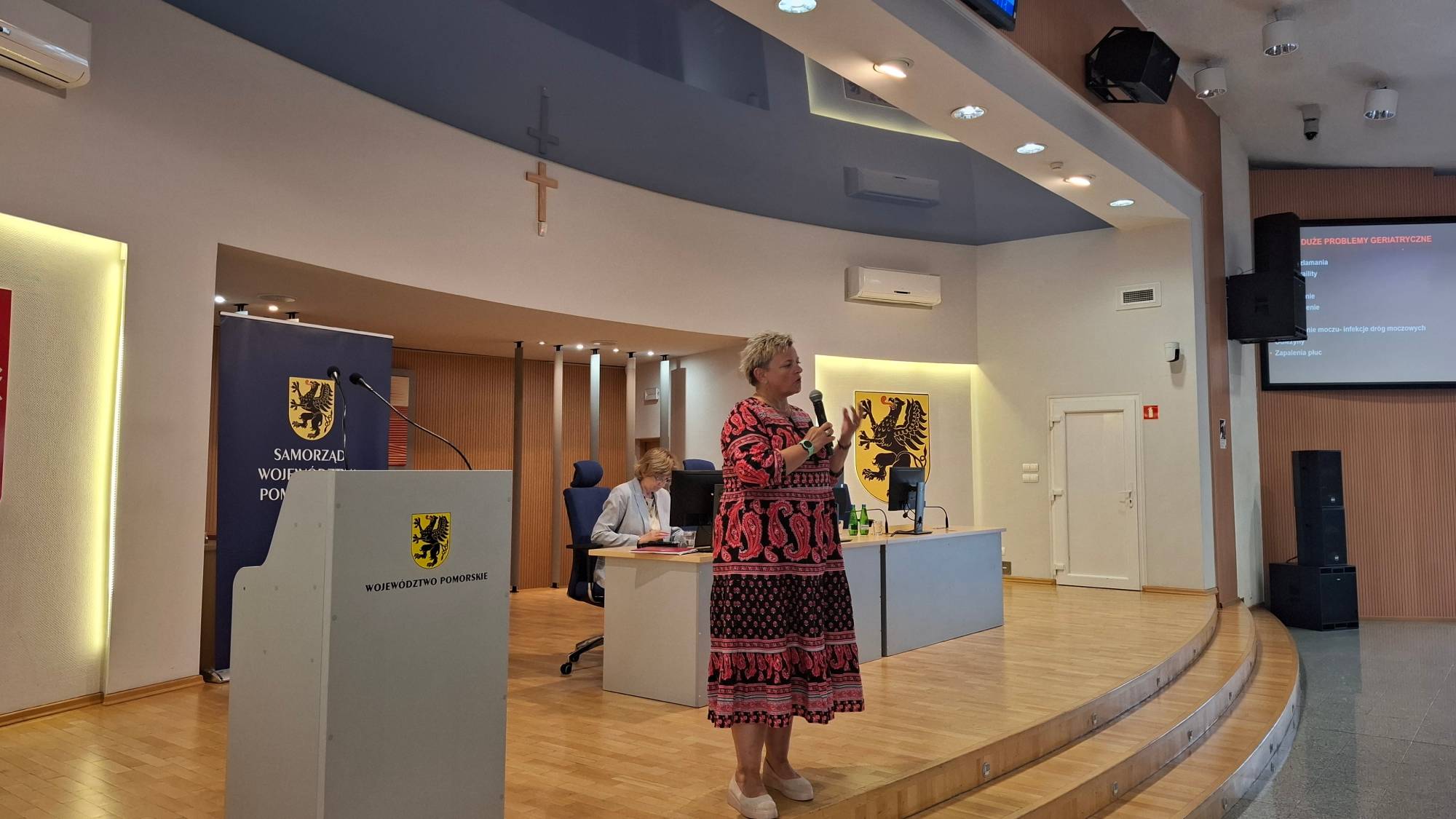 Kobieta w długiej, wzorzystej sukni przemawia do zgromadzonych, trzymając w ręku mikrofon i gestykulując. Stoi na podwyższeniu w sali konferencyjnej, przed mównicą z herbem województwa pomorskiego. W tle widoczne są godła regionu, ekran z prezentacją oraz siedząca przy stole inna uczestnica spotkania. Wykład dr Dagmary Wiewiórkowskiej-Garczewskiej w czasie konferencji dla seniorów w Gdańsku