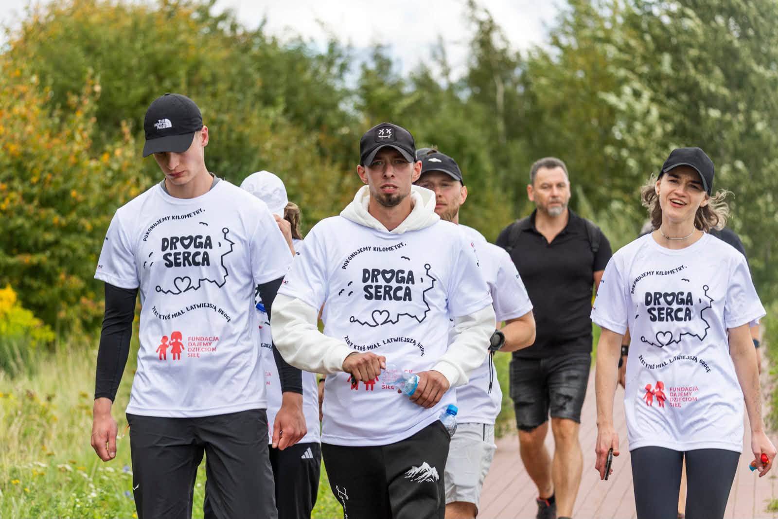 Grupa uczestników charytatywnego marszu "Droga Serca" w białych koszulkach z logo akcji, idąca ścieżką w plenerze