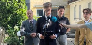 Czterej mężczyźni stojący przed budynkiem, w tym młody działacz Młodzieży Wszechpolskiej w garniturze czytający oświadczenie z kartki przed mikrofonami radiowymi, wspierany przez kolegów i polityka Konfederacji