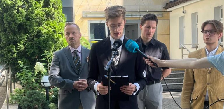 Czterej mężczyźni stojący przed budynkiem, w tym młody działacz Młodzieży Wszechpolskiej w garniturze czytający oświadczenie z kartki przed mikrofonami radiowymi, wspierany przez kolegów i polityka Konfederacji