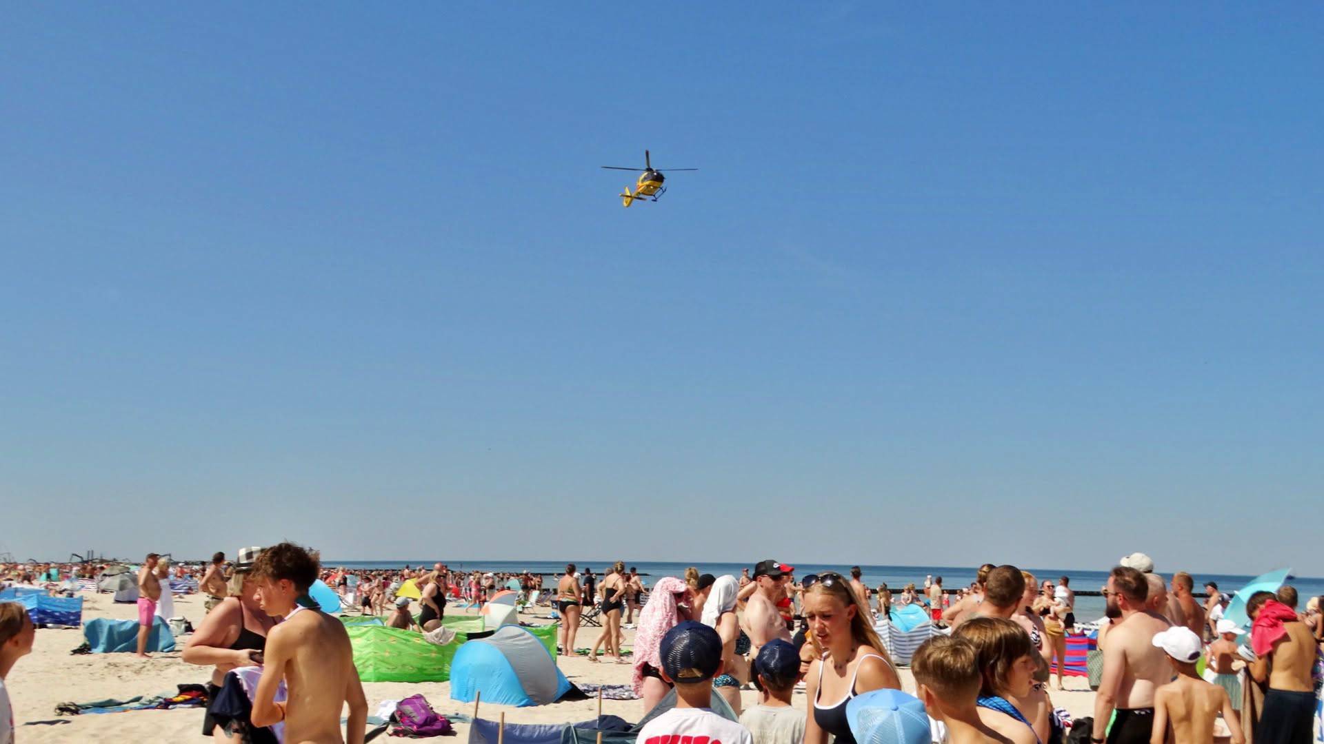 Akcja ratunkowa na plaży w Ustce, gdzie tonęło dziecko. Żółty śmigłowiec Lotniczego Pogotowia Ratunkowego leci nisko na tle bezchmurnego nieba nad zatłoczoną, piaszczystą plażą z parawanami i wypoczywającymi turystami w Ustce