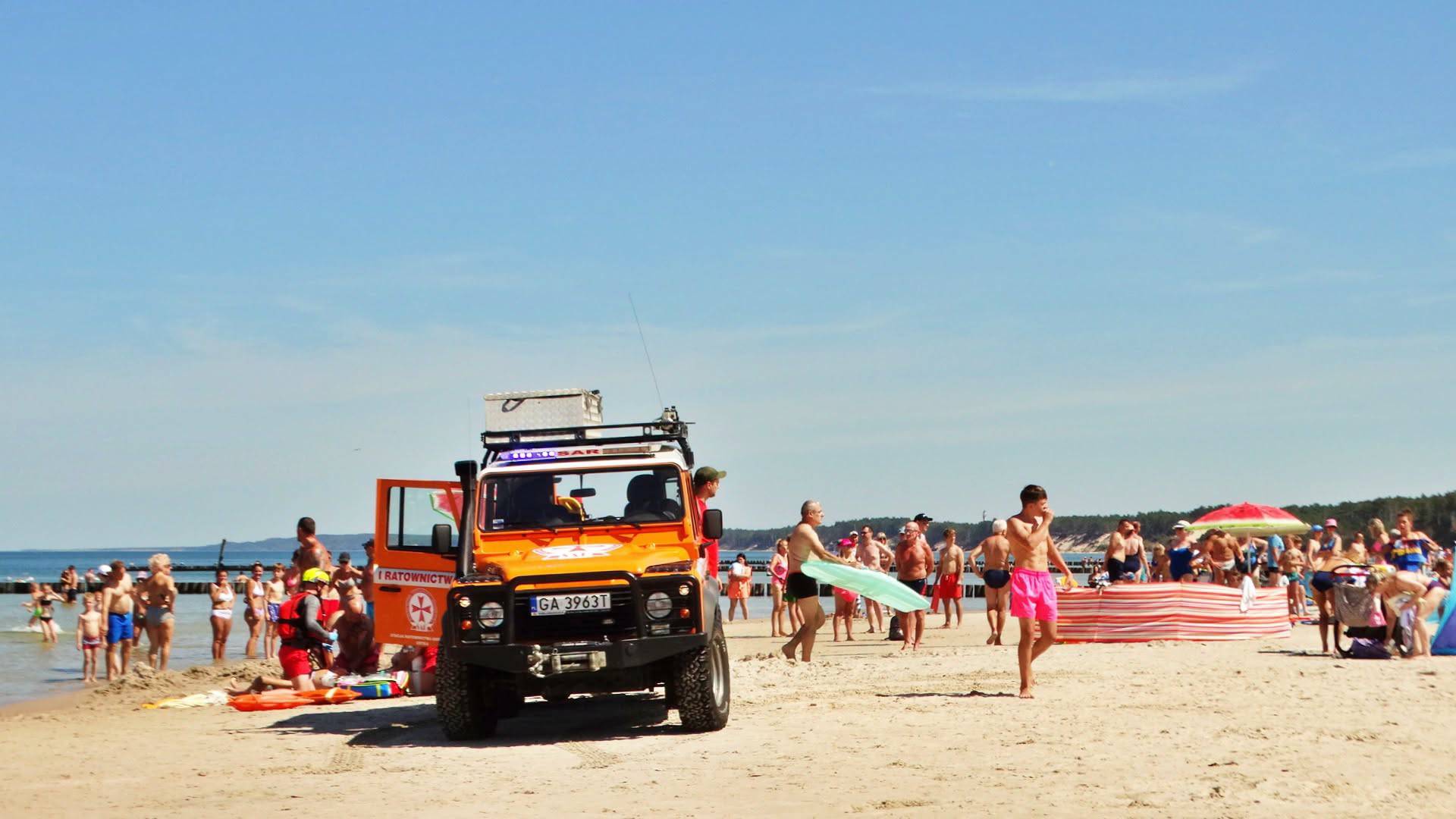 Akcja ratunkowa na plaży w Ustce, gdzie tonęło dziecko. Pomarańczowo-biały pojazd terenowy ratowników z widocznym numerem rejestracyjnym GA 3963T stoi na piaszczystej plaży w Ustce. Wokół pojazdu i na tle morza widać licznych plażowiczów oraz ratowników udzielających pomocy