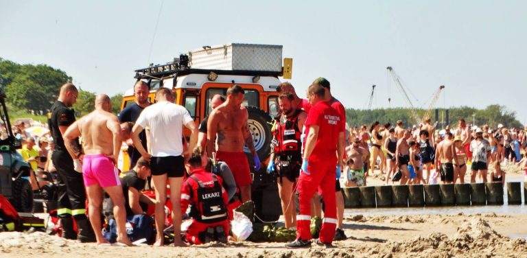 Akcja ratunkowa na plaży w Ustce, gdzie tonęło dziecko. Grupa ratowników w czerwonych i czarnych strojach oraz strażacy stoją na piaszczystej plaży wokół pacjenta udzielając pierwszej pomocy. W tle widoczny jest specjalistyczny ratowniczy pojazd terenowy, a w dalszym planie tłum plażowiczów nad brzegiem morza i drewniane falochrony