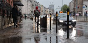 Mokra ulica w Gdańsku podczas deszczu. Na pierwszym planie widoczne są czarne słupki ostrzegawcze i mokry, odbijający światło chodnik. W tle jadący rowerzysta w czerwonej kurtce z kapturem oraz przejeżdżający czarny samochód, spod którego kół pryska woda. IMGW ostrzega przed burzami i deszczem na Pomorzu
