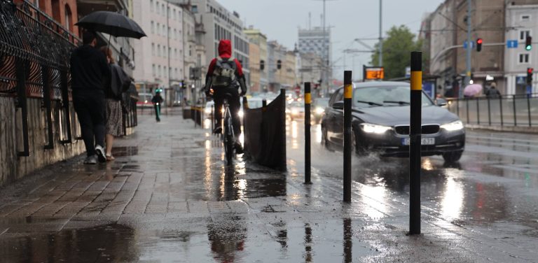 Mokra ulica w Gdańsku podczas deszczu. Na pierwszym planie widoczne są czarne słupki ostrzegawcze i mokry, odbijający światło chodnik. W tle jadący rowerzysta w czerwonej kurtce z kapturem oraz przejeżdżający czarny samochód, spod którego kół pryska woda. IMGW ostrzega przed burzami i deszczem na Pomorzu
