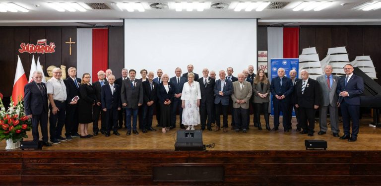 Grupa kilkudziesięciu osób - głównie działaczy dawnej opozycji antykomunistycznej oraz przedstawicieli Instytutu Pamięci Narodowej - pozuje do pamiątkowego zdjęcia na scenie w historycznej Sali BHP Stoczni Gdańskiej, gdzie odbyła się uroczystość wręczenia Krzyży Wolności i Solidarności W tle widoczne są polskie flagi, krzyż, logo "Solidarności" oraz ekrany projekcyjne