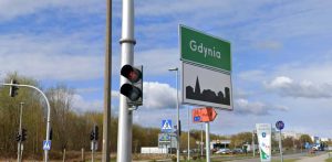 Zielona tablica drogowa z białym napisem "Gdynia" oraz znak informujący o wjeździe w obszar zabudowany, ustawione przy drodze obok sygnalizatora świetlnego