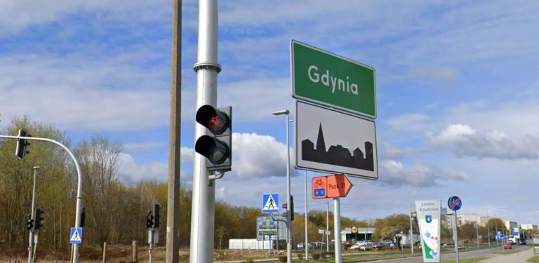 Zielona tablica drogowa z białym napisem "Gdynia" oraz znak informujący o wjeździe w obszar zabudowany, ustawione przy drodze obok sygnalizatora świetlnego