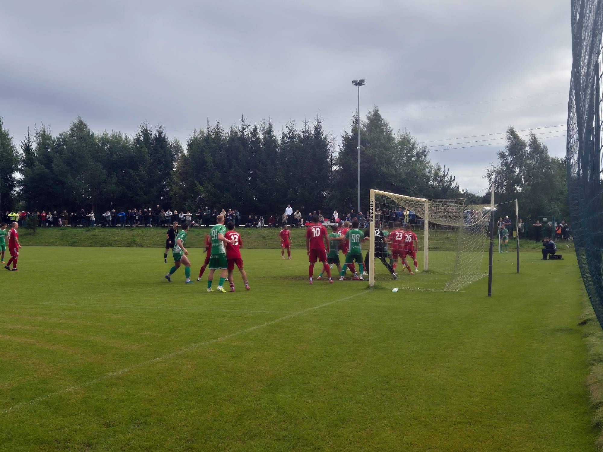 Mecz 4. kolejki 4. pomorskiej ligi piłkarskiej pomiędzy drużyną Anioły Garczegorze a Gryfem Słupsk. Zdjęcie przedstawia fragment meczu piłkarskiego na zielonej murawie. W polu karnym gromadzą się zawodnicy drużyn przeciwne w czerwonych i zielonych koszulkach oraz bramkarz w czarnym stroju, walczący o piłkę pod bramką. W tle widoczny jest szpaler kibiców stojących wzdłuż boiska oraz wysoki las iglasty pod pochmurnym niebem