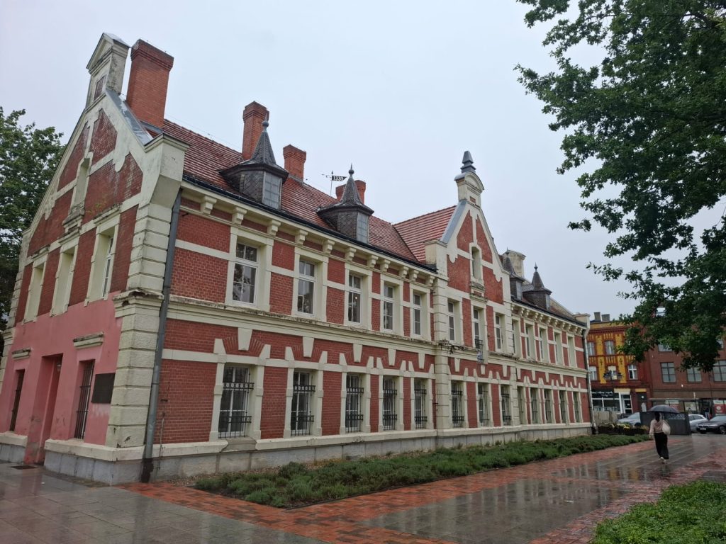 Dwupiętrowy, zabytkowy budynek z czerwonej cegły o neorenesansowej architekturze, z ozdobnymi wieżyczkami i białymi detalami na elewacji. Przed obiektem ciągnie się pas zieleni oraz mokry od deszczu chodnik, po którym idzie osoba z parasolem