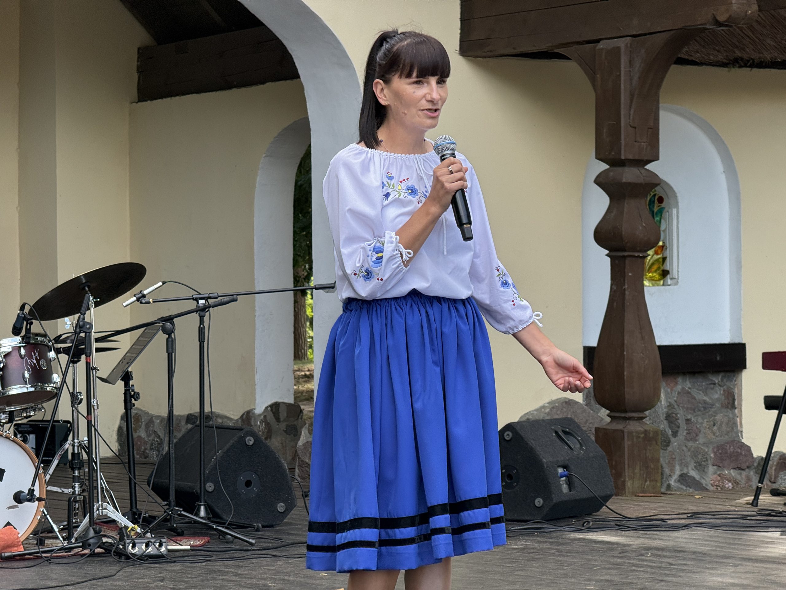 Kobieta o ciemnych włosach upiętych w kucyk stoi na scenie plenerowej z mikrofonem w ręku i przemawia (Olga Kuklińska, zdobywczyni Grand Prix 49. Wielewskiego Turnieju Gawędziarzy). Ubrana jest w białą tradycyjną bluzkę z haftem kaszubskim na piersi i rękawach oraz niebieską rozkloszowaną spódnicę do kolan z dwoma czarnymi paskami przy dolnym brzegu. W tle widać drewnianą konstrukcję sceny, zestaw perkusyjny i głośniki odsłuchowe