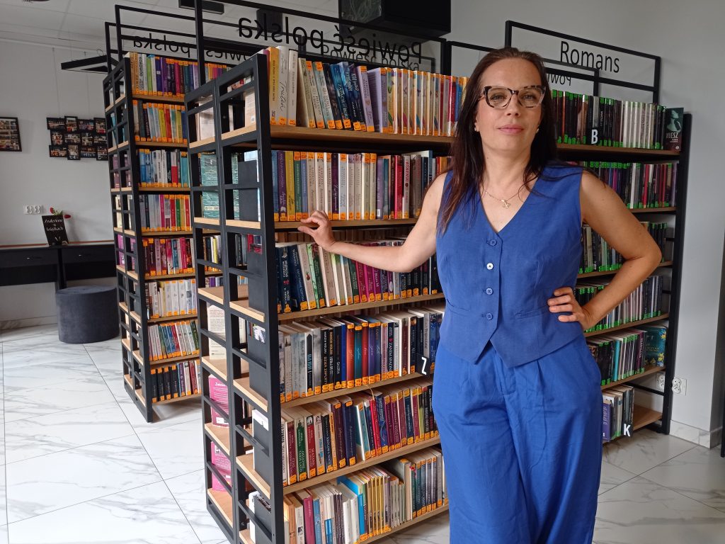 Na zdjęciu Anna Zakrzewska, kierowniczka Biblioteki Ratuszowej, na tle regałów z kolorowymi książkami