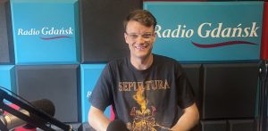 Jakub Mielewczyk, uśmiechnięty mężczyzna w okularach i czarnym t-shircie z logo zespołu Sepultura, siedzi w studiu nagraniowym przy mikrofonie. W tle widać niebiesko-szare panele akustyczne z logo Radia Gdańsk. Gość poprowadzi zajęcia teatralne w Nowym Teatrze im. Witkacego w Słupsku