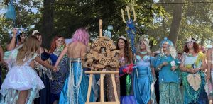 Na zdjęciu widzimy 12 kobiet na scenie, przebranych w suknie przypominające syreny. Na scenie stoi także sztaluga z wykonanym na drewnie napisem dożynki rybne.