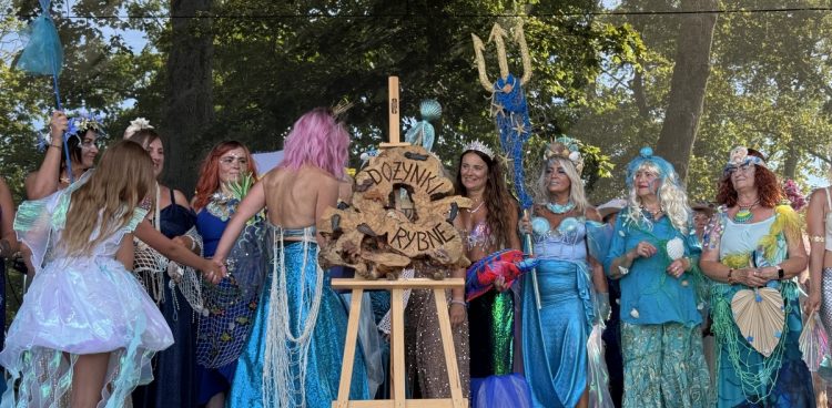 Na zdjęciu widzimy 12 kobiet na scenie, przebranych w suknie przypominające syreny. Na scenie stoi także sztaluga z wykonanym na drewnie napisem dożynki rybne.