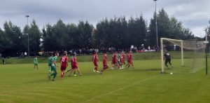Mecz 4. kolejki 4. pomorskiej ligi piłkarskiej pomiędzy drużyną Anioły Garczegorze a Gryfem Słupsk zakończony remisem 1:1. W polu karnym widoczni są zawodnicy w czerwonych i zielonych strojach oraz bramkarz w czarnym ubraniu. W powietrzu nad boiskiem leci piłka, a w tle zgromadzona jest publiczność oraz ściana lasu