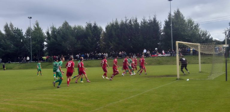 Mecz 4. kolejki 4. pomorskiej ligi piłkarskiej pomiędzy drużyną Anioły Garczegorze a Gryfem Słupsk zakończony remisem 1:1. W polu karnym widoczni są zawodnicy w czerwonych i zielonych strojach oraz bramkarz w czarnym ubraniu. W powietrzu nad boiskiem leci piłka, a w tle zgromadzona jest publiczność oraz ściana lasu