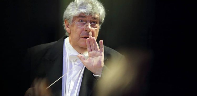 Dyrygent Jan Łukaszewski we fraku i okularach, trzymający batutę i wykonujący gest otwartą dłonią