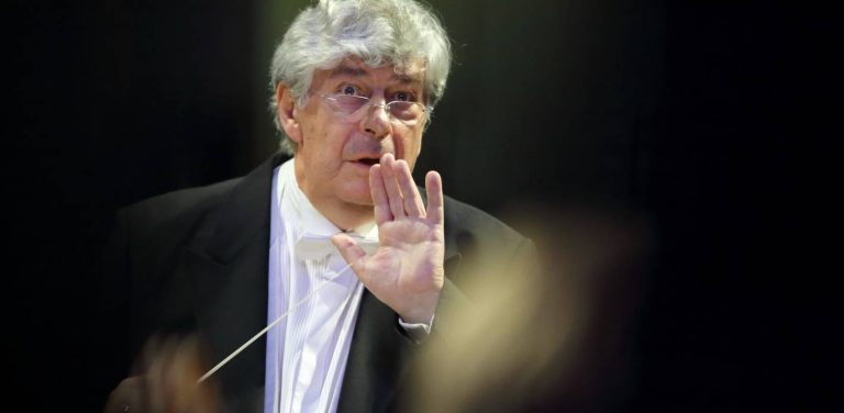 Dyrygent Jan Łukaszewski we fraku i okularach, trzymający batutę i wykonujący gest otwartą dłonią