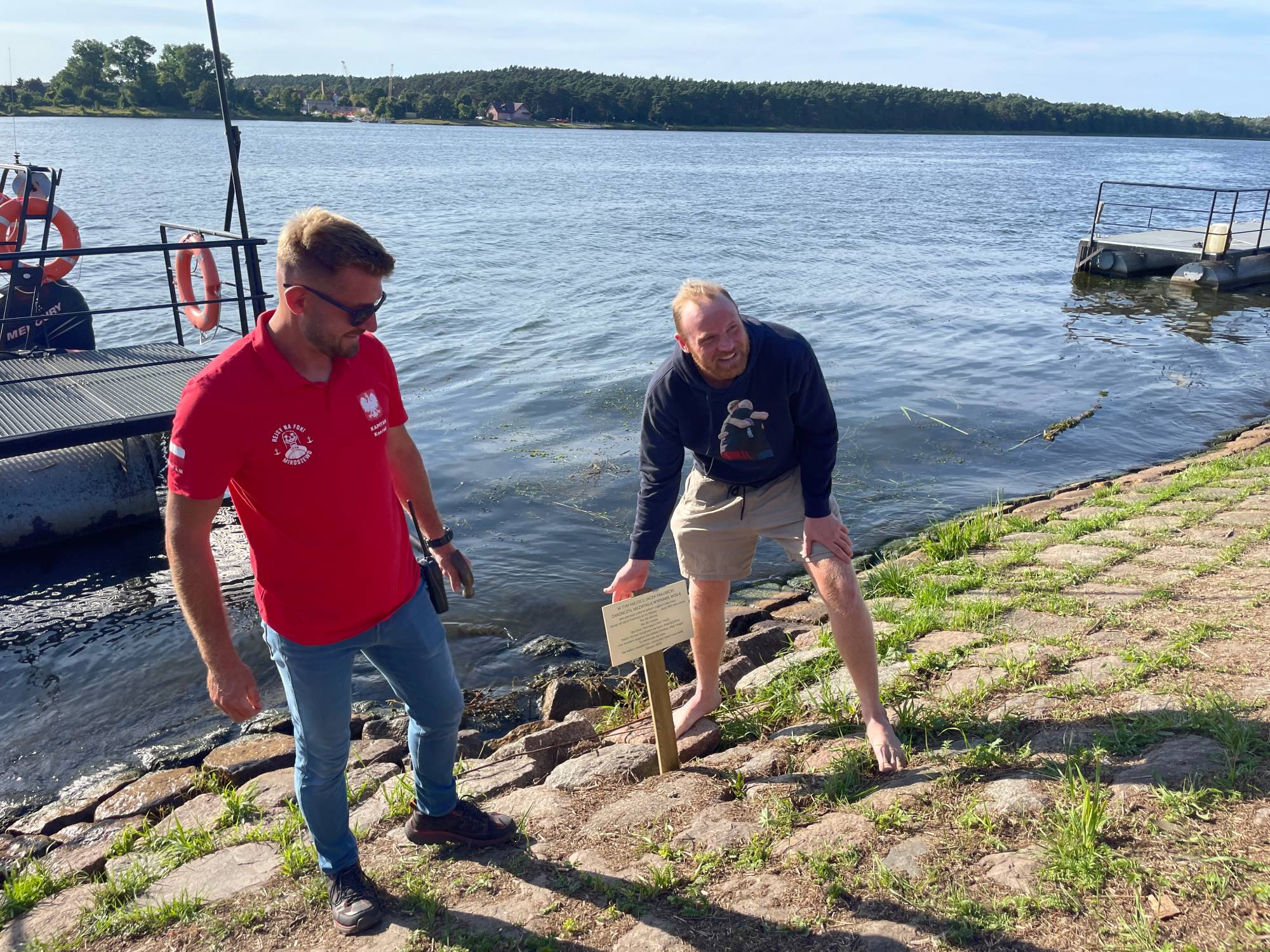 Bosy Jacek Pałubicki w bluzie i spodenkach pochyla się nad pamiątkową tabliczką wbitą w kamienisty brzeg rzeki. Obok niego stoi mężczyzna w czerwonej koszulce polo, a w tle widać Wisłę i pomost. Mieszkaniec Rumi przepłynął wpław Wisłę (942 km), zbierając pieniądze dla dzieci z alkoholowym zespołem płodowym