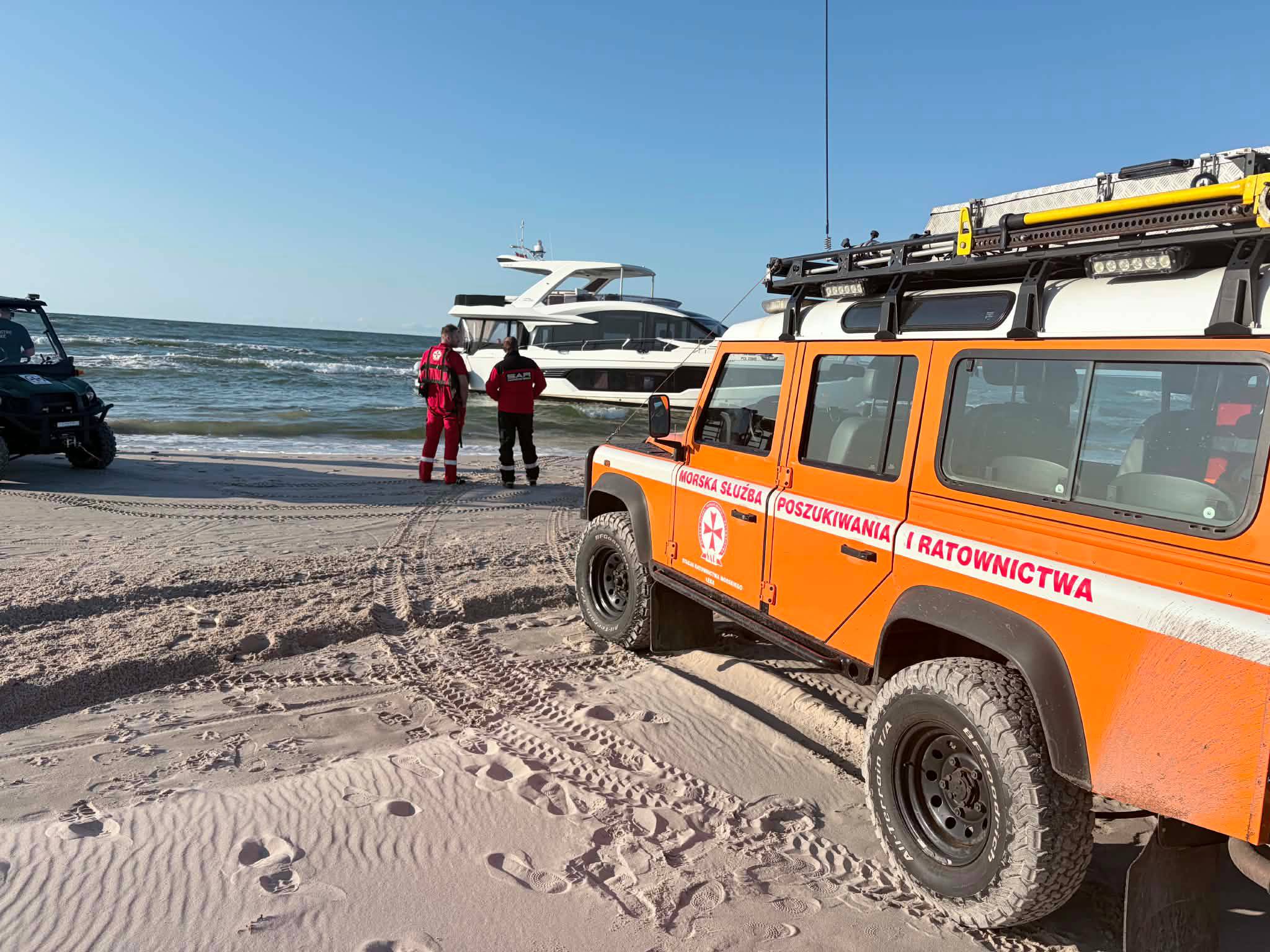 Pomarańczowy samochód terenowy Morskiej Służby Poszukiwania i Ratownictwa (SAR) zaparkowany na piaszczystej plaży. Dwóch ratowników w czerwonych kombinezonach stoi przy brzegu i obserwuje biały jacht motorowy osiadły na płyciźnie