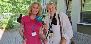 Joanna Rutkowska trzymająca w dłoniach kije do nordic walking oraz Anna Rębas. W tle fragment budynku i zielone drzewa